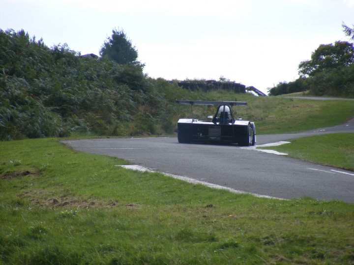 Loton Park Pistonheads Bhc - The image shows a green and grass-covered park or natural area with a paved road. A quad bike with a windshield and harness points for a sidecar is seen with the sidecar empty, and it is maneuvering on the road. A hedge marks the boundary between the road and the green area, and throughout the scene, there are several trees, providing a sense of the outdoors and a peaceful setting. The sky is overcast, suggesting a cloudy day. The overall atmosphere of the image is serene and natural.