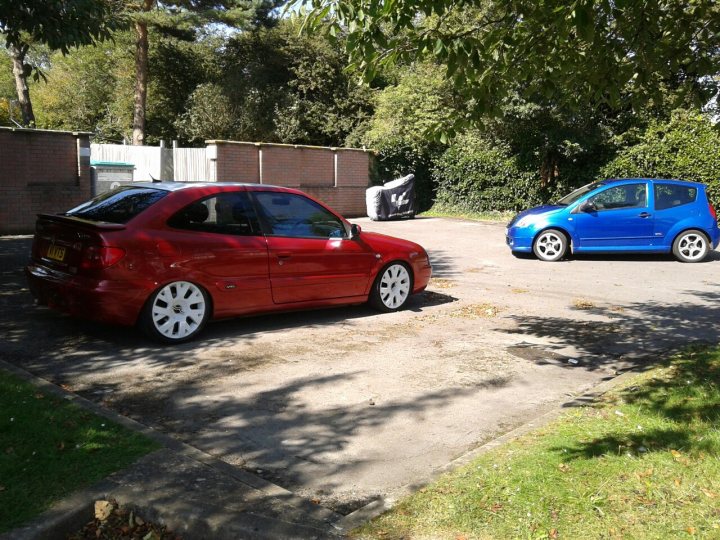 My Two French beauties. - Page 1 - French Bred - PistonHeads - The image captures a scene with two small hatchback cars parked in a narrow, curving space within a driveway. The car closer to the camera has a vibrant red hue, while the one farther away appears in a rich blue color. Both vehicles have black and silver accents. They are parked between grassy areas and a fence, with trees visible in the background.