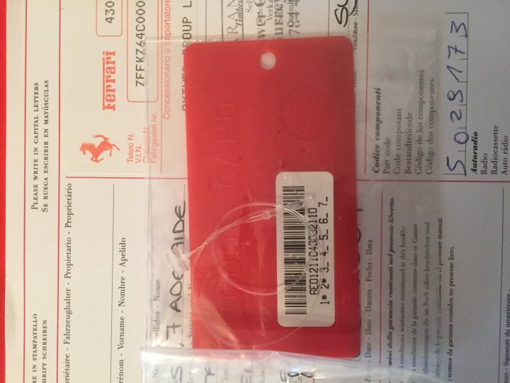 Stereo code lost HELP Please  - Page 1 - Ferrari V8 - PistonHeads - The image presents a close-up view of a red identification card lying on a bed of paper. The card commands attention due to its large size. It features a barcode in the center, a common feature in many types of identification cards. Complementing the barcode, there's a space indicated for the photograph of the owner, yet the photo is missing from the card. The card also has text elements, including a visible text line, where perhaps the name or identification number would typically be printed, although these texts are not clearly legible in this image.