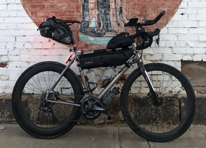 Tail light for seat stay - Page 2 - Pedal Powered - PistonHeads - The image presents a scene featuring a robust, dark-colored bicycle leaning against a richly colored brick wall. The bicycle is equipped with prominent dark-colored wheels and a chain case attached to the back tire. The handlebars are slightly turned, possibly signifying that someone just rode it and left it temporarily. The rear of the bicycle is adorned with a backpack, possibly containing essential items. Notably, the brick wall is not plain; it features a charming mural of a bird, adding a touch of whimsy to the otherwise urban setting.