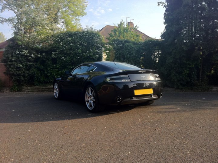 Changed Pistonheads Spot - The image shows a modern black car parked on a paved driveway. The vehicle is sleek and has a yellow license plate mounted in the center of its rear bumper. To the left of the car, there's a neatly trimmed hedge, and to the right, a privacy fence which is common for residential properties. The natural light and clear blue sky suggest it's a sunny day. The car is the focal point of the image, and there is no text or other objects in the frame.