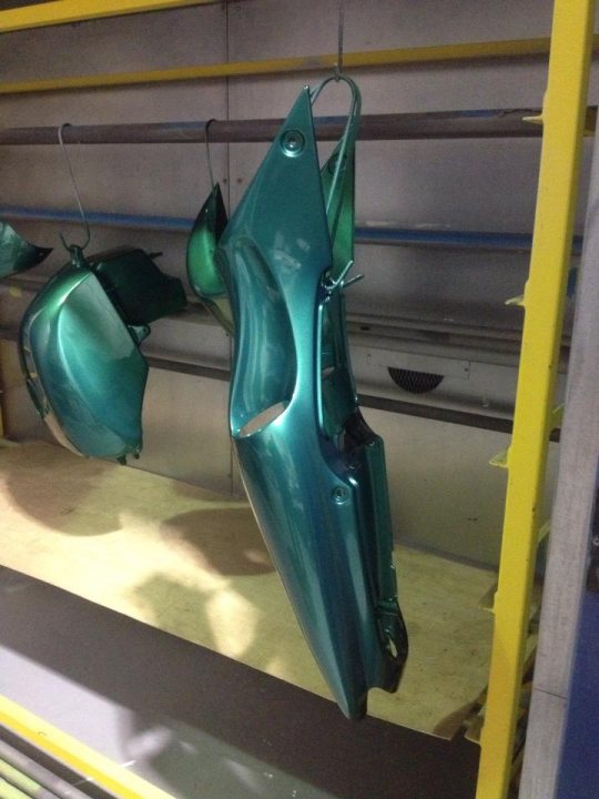 A pair of scissors sitting on top of a table - Pistonheads - The image shows a vibrant teal-colored car part hanging on a hook from a rack. The part appears to be a fender or bumper, with an intricate design and visible screws along its edges. The background suggests it's within the confines of a garage or workshop, with white walls and a yellow and grey support shelf. There's another fender or bumper directly below it, also hanging from a hook, indicating that this may be a storage area for car parts.