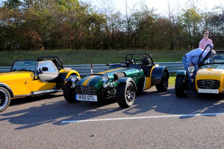 PH SS Silverstone Pictures & Video - Page 1 - Caterham - PistonHeads - This image depicts a scene of four vintage cars parked on what appears to be a roadside or race track, with greenery in the background. The cars, all convertible or roadster models, are painted in bright, vibrant colors: yellow, green, and black. They each have distinct vintage designs and are given the distinguishing model names "R50 OTL," "R7 KCL," and "R7 SNU," as indicated on the vehicles. The sky is visible in a clear blue, indicating it's a bright and sunny day.