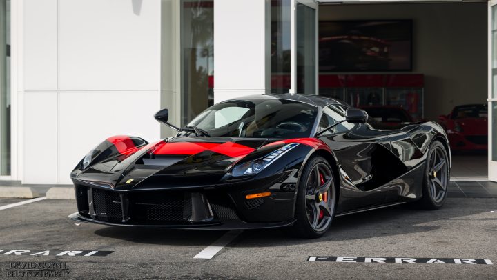 LaFerrari - just delivered. Black. - Page 1 - Ferrari V12 - PistonHeads - The image displays a sleek, black sports car parked in front of a building with a prominent door. The car is parked neatly on the far side of a marked parking space, with its design and color drawing attention to its speed and style. The reflection of the car on the shiny surface next to it implies that the photo might have been taken on a sunny day, underlining the car's distinctive appearance. The placement of Ferrari branding suggests that this car is a model of the luxury automobile brand.