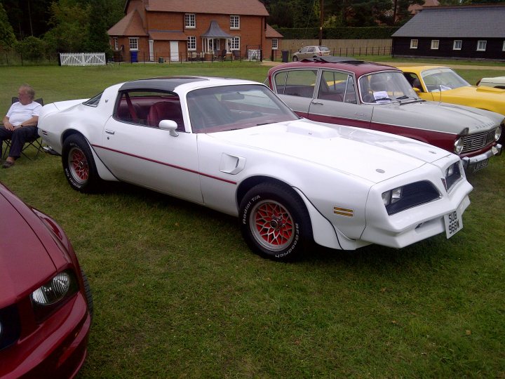 Pistonheads - The image features a classic white muscle car parked on a grass field. The car has a prominent front grille and bold red and white stripes down its side and through the center of the rear wing. To the right, there is a vintage red and white striped European sedan. Further to the far right, you can see the back of an older, yellow compact car. In the background, there's a rustic wooden building with a dirt driveway, suggesting a rural or countryside setting. An elderly person is seated near the cars, partially visible in the left side of the picture.