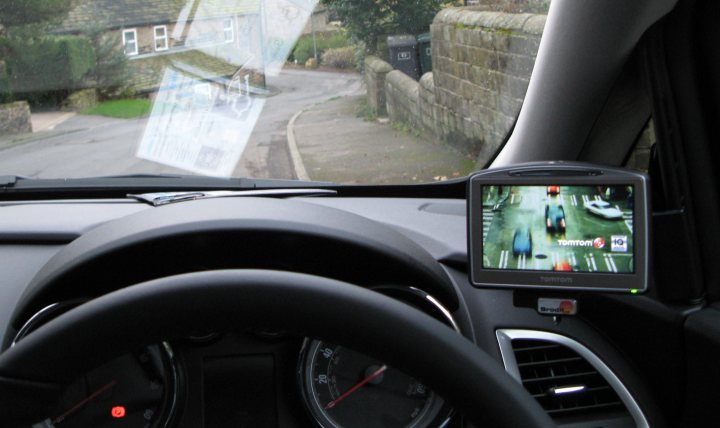 Pistonheads - The image shows the interior of a vehicle, taken from the perspective of the driver. The dashboard is visible, displaying the instrument cluster with the speedometer and tachometer, as well as various gauges, dials, and buttons. On the right side of the dashboard, there is a screen mounted on a base displaying a GPS or navigation system, which shows a visual representation of a route with traffic information at the bottom of the screen. Through the windshield, one can see a residential street with a car in motion, driving past a stone wall and hedges.