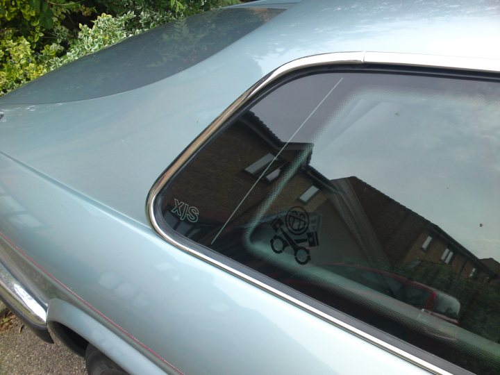 Pistonheads - The image shows a close-up of a car's rear windshield with a sticker depicting a smiling face on the window. The windshield is framed by the car's metallic side mirror. In reflection, a brick wall can be seen behind the car. The view is taken from an angle that partially obscures the back of the car. There are some green foliage in the background. The overall color tones are muted, suggesting an overcast or cloudy day.