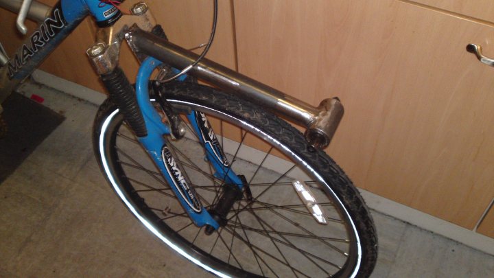 Pistonheads - This image features a close-up of a blue bicycle wheel with a thick black tire. The frame of the bike is partially visible in the top left corner, suggesting that the photo was taken with the bike upside down. The wood-grained texture of a cabinet or panel is visible on the right, indicating that the bicycle is likely indoors. There is a lock attached to the chain ring and rear wheel hub, which is a common anti-theft measure for bicycles.