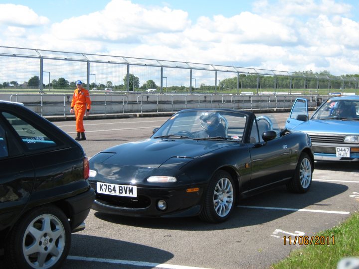 Pistonheads - The image captures a scene at a parking lot. A black sports car is parked in the foreground, extending into the scene on both sides. The vehicle is sleek and modern, with a design that suggests speed and power. Behind the sports car, there are other cars and a fence. Beside it stands an individual, dressed in an orange tracksuit, offering a striking contrast of colors to the dark car. This person appears to be engaged in an activity related to the cars. The setting, combined with the activities, suggests a possible race or car show event.