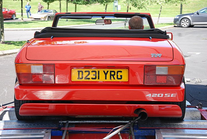 Auto electrician. - Page 1 - South Wales - PistonHeads - The image shows a bright red MGB car being towed by a yellow tow truck. The car is in the drop position, and the tow truck is not in any specific motion. There are visible straps connecting the car to the tow truck. The scene takes place on a street with some surrounding cars and individuals.