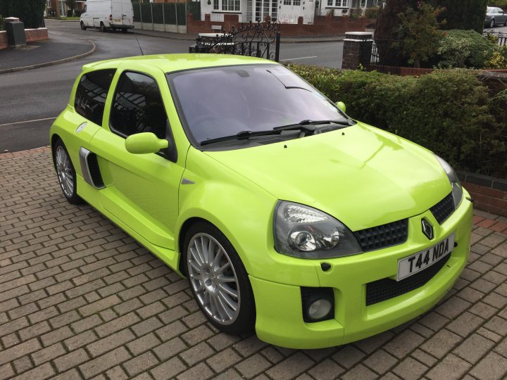 RE: Renault Clio V6: Spotted - Page 1 - General Gassing - PistonHeads - This image features a vibrant lime green car parked on a brick driveway adjacent to a street. The car appears to be a compact vehicle with a distinctive bright color, which gives it a sporty and unique look. The body of the car has a reflective sheen to it, suggesting a glossy finish. Passenger windows and the windshield are tinted or darkened for privacy, and there are visible features such as wheel rims and door handles. In the background, there are residential buildings and a partial view of a bus parked on the road.