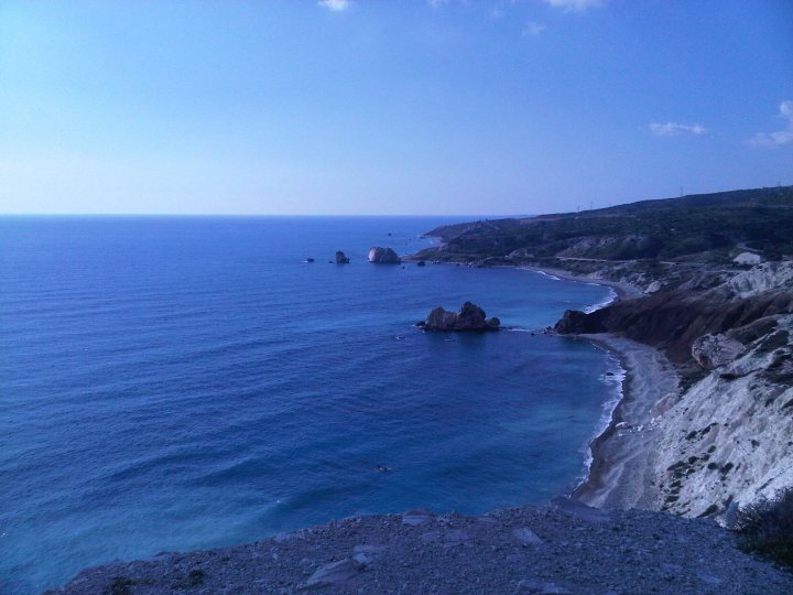 Hello from Cyprus! - Page 1 - Aston Martin - PistonHeads - The image captures a breathtaking coastal scene. The ocean, painted in a deep blue color, stretches out to meet the clear, cloudless sky. A few small islands can be seen in the distance, dotting the horizon with their presence. On the left side of the image, a cliff face drops steeply towards a small beach, which is partially covered in sand. The surreal colors and the serene atmosphere make this image a picturesque representation of a coastal landscape.