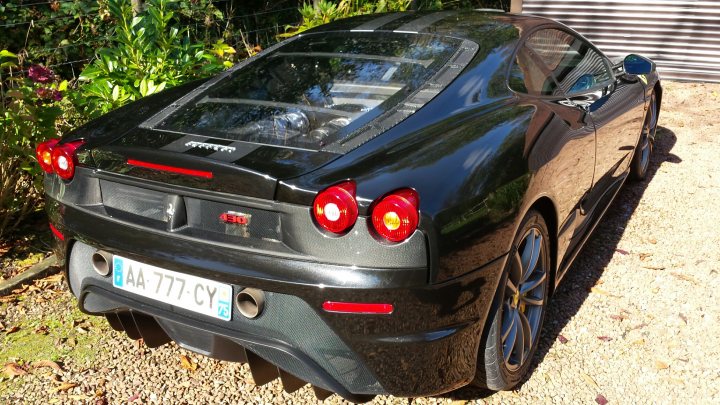 New Scuderia Owner - What a car!!! - Page 1 - Ferrari V8 - PistonHeads - The image shows a luxurious black sports car parked on a gravel surface, casting a shadow on the ground to its right. It has a distinctive design, with angular body lines and an aerodynamic front bumper. The car features a rear spoiler, and the side badge reads "Scuderia." There is also a wing on the hood that says "Cyber TwinTurbo." Light is reflecting off its glossy paint, and there's an impression of the car's music system business sticker underneath the rear window. The setting appears to be outdoors, perhaps in a garden or driveway, with greenery visible in the background.