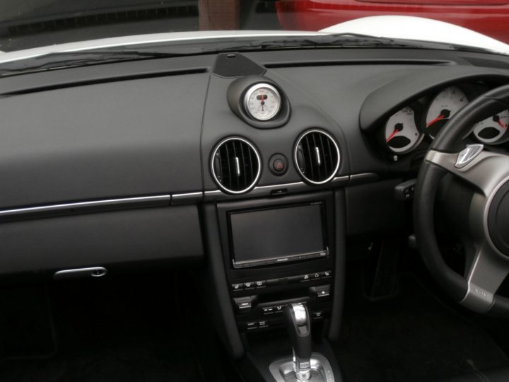 Cayman 997.2 Radio Options - Page 1 - Boxster/Cayman - PistonHeads - The image features the interior of a car, focusing on the driver's side. The dashboard is clearly visible with controls for the radio, climate control, and other vehicle functions. A digital display screen is located just below the main dashboard, suggesting a feature-rich entertainment system. There is a red car visible through the window on the right, which contrasts with the dark interior of the current vehicle. The car appears to be neatly parked in a secure location, possibly a closed parking structure or a sheltered area.