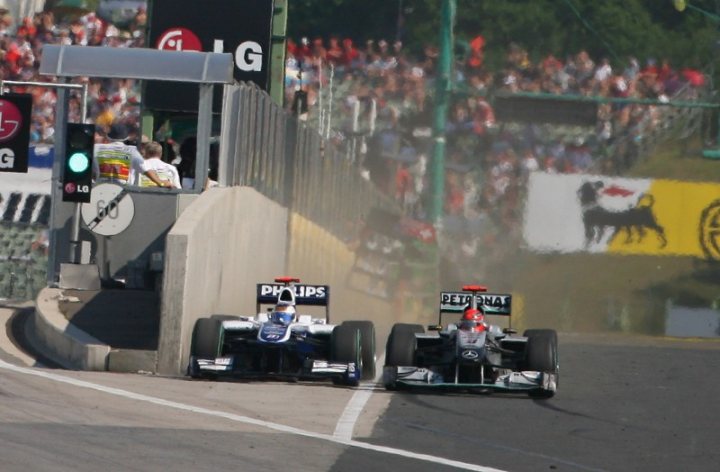 How many races until Schumacher has/does one of his "stunts" - Page 23 - General Motorsport - PistonHeads - This image captures a thrilling moment in a Formula One race. Two racing cars, painted in vibrant shades of red and silver, are in the midst of an intense battle with a dust trail flung up behind them. The cars are racing on a track, the blue lining of which stands out against the surrounding landscape. The atmosphere is electric, with the stands teeming with spectators, each engrossed in the unfolding action on the track. The name "Petrusana" and the logo of the "LG Principal sponsor" are prominently displayed, indicating their involvement in the race. The cars are adorned with red numbers, adding another layer of complexity to the scene. This image encapsulates the speed, intensity, and excitement inherent to Formula One racing.