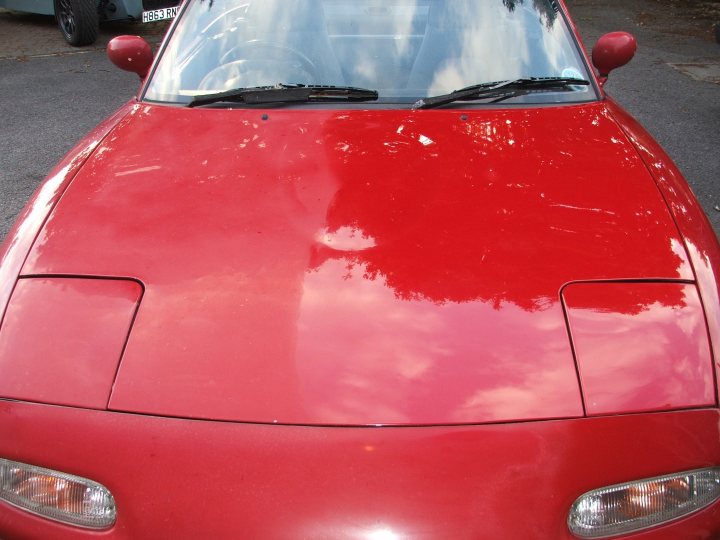 Pistonheads Meguiars - The image features a vibrant red sports car, parked on a concrete surface with trees and foliage in the background. The car's shiny body reflects the light, enhancing its sleek design. The windows and the open driver's side door suggest the car is ready to be driven, while the presence of another car and road markings indicate this is a public parking area.