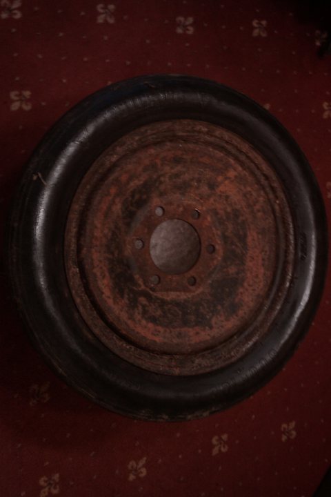 Sized Odd Pistonheads Tyre - The image shows a large brown rubber tire lying flat on a surface with a patterned red and yellow carpet beneath it. The tire appears to be old and worn, with rust and dirt accumulated on its tread, and the metal hub in the center is visible and looks similarly corroded. The tire takes up most of the frame, suggesting it's the main focus of the image.