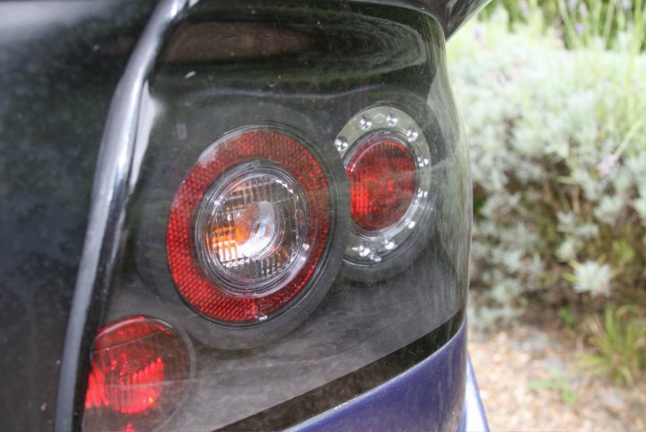 Noble Lights Deffusers Pistonheads Led - The image shows a close-up view of the circular red and orange tail lights of a vehicle. These particular red taillights have a distinct orange chevron design at their center. The lights are set into a structure that appears to be made of paint or a material with a reflective finish. In the background, there are blurred green plants, providing a natural context to the vehicle. The image seems to have been taken outdoors, given the presence of natural elements.