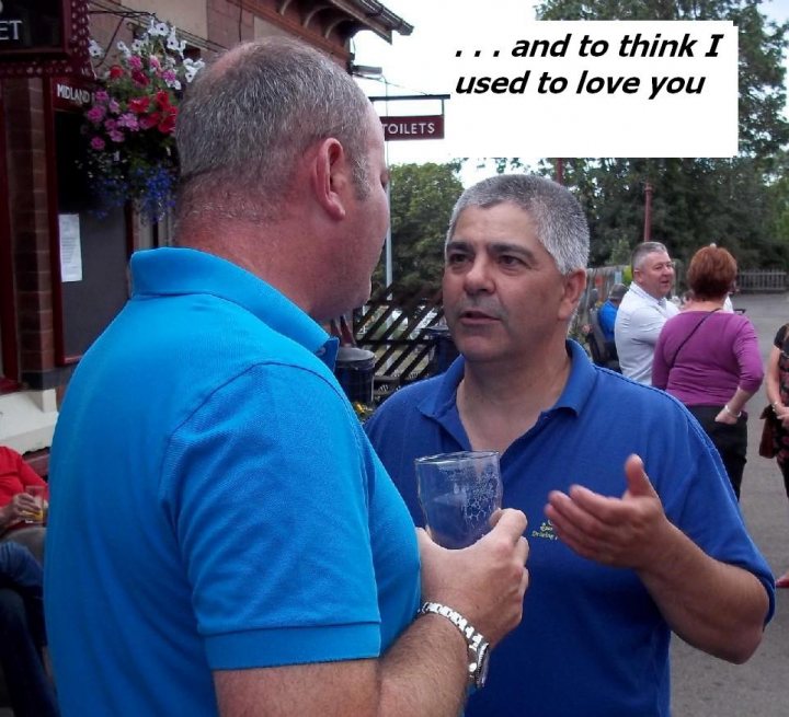 Run out this sunday - Page 1 - Northamptonshire - PistonHeads - The image is a candid photograph featuring two adult males in outdoor daylight. The man on the left is bald and wearing a light blue polo shirt. He has a short beard and is holding a glass of beer. The man on the right has gray hair and is in the middle of speaking or explaining something to the other man. He is wearing a dark blue polo shirt. Both men appear to be engaged in a casual conversation or interaction. In the background, there are potted plants, a sign that reads "MEN'S LOUNGE," and other individuals that are not the focus of the picture.