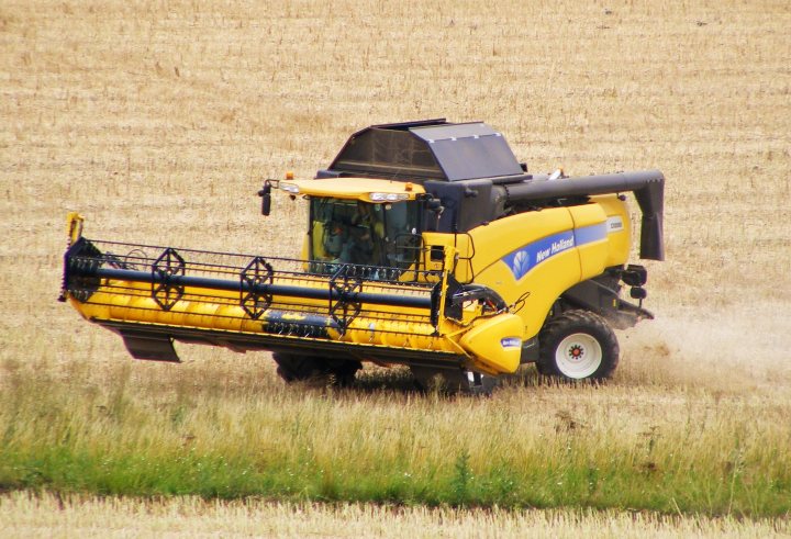 RE: Mr Big Stuff: Claas Lexion 530 Combine - Page 4 - General Gassing - PistonHeads - This is a color photograph of a yellow agricultural tractor in the process of cultivating a field. The tractor is seen from a side angle, revealing the machinery and its current operation, as evidenced by the cloud of dust or soil being pushed up from the earthy surface of the field. The field extends into the distance, marked by a few interspersed bushes, indicating a period of plant growth that is not yet fully tilled. There are no visible texts or brands in the image.