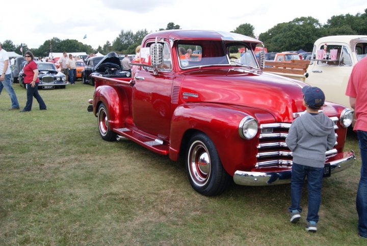 Anyone going to the NASC 40th Nats at Trinity Park? - Page 1 - East Anglia - PistonHeads - In the image, a vibrant red classic pickup truck is parked on a grass field, surrounded by various cars and people. The pickup truck, with its glossy finish, is the central focus. A child and a man are standing next to the truck, possibly admiring its design or features. The truck is not just parked but also taken for a spin, as indicated by its partially raised hood. The setting appears to be an outdoor car show, where enthusiasts gather to showcase their vintage machines.