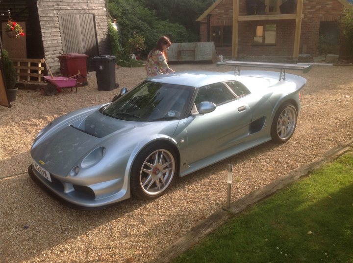 Another newbie - Page 1 - Noble - PistonHeads - The image showcases a sleek silver sports car parked in the front yard of a house. The car's prominent rear spoiler and streamlined coupe lines are indicative of its design, suggesting it's possibly a Lamborghini model. The driveway is well-maintained, indicating a residential setting. A person can be seen sitting on the car, conveying a sense of relaxation and enjoyment in the pleasant surroundings.