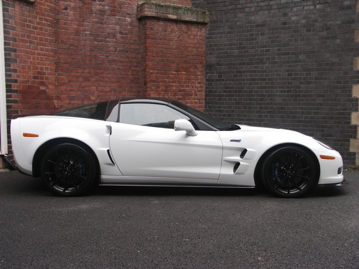 How about some ZR-1 photos to cheer you up? - Page 1 - Corvettes - PistonHeads - The image shows a white sports car parked on what appears to be a road or pavement, set against a brick wall with a building adjacent to the car. The car's design suggests it's a convertible model with a sleek and modern aesthetic. The doors are closed, indicating that the car is not currently in use. The environment looks urban or suburban, with the pavement surface indicating recent maintenance for the area.