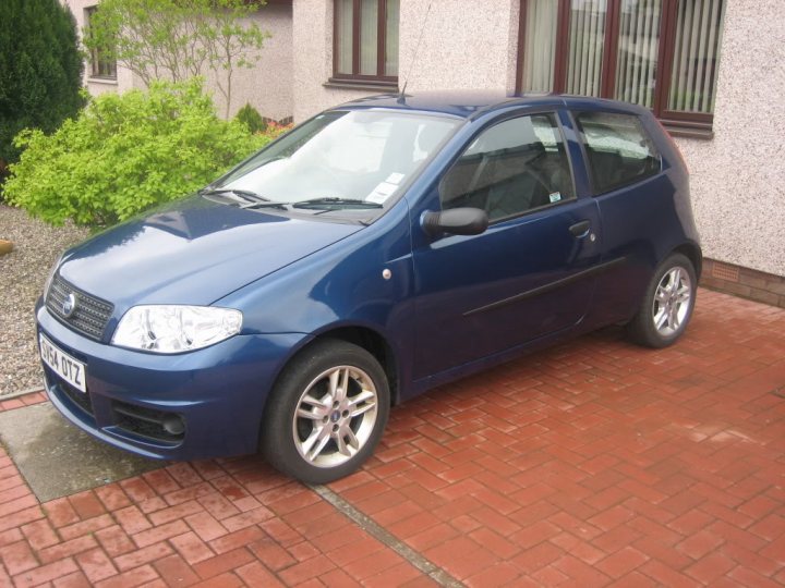 My first car! - Page 1 - Readers' Cars - PistonHeads - The image showcases a small, dark-colored car parked on a brick driveway. The vehicle has a shiny, well-groomed appearance and is positioned at a slight angle to the camera. The car features a sunroof and alloy wheels, adding to its sleek look. In the background, there is a building with a partial view of a person walking near a window. The clouds in the sky suggest that it is a daytime scene.