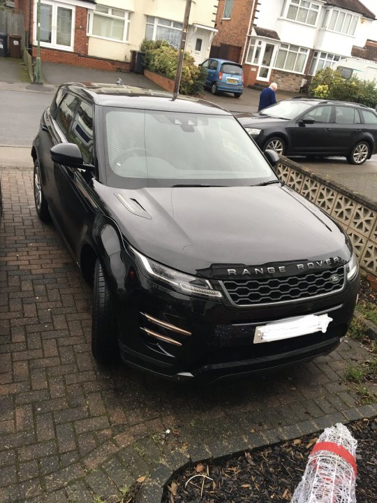 A car is parked on the side of the road - The image shows a modern black SUV parked on the side of a street. The vehicle is parked in front of a brick wall and near a driveway with a car in it. There are no people visible in the immediate vicinity, but some cars can be seen further down the street. The surroundings suggest a residential area with houses and driveways on both sides of the road.