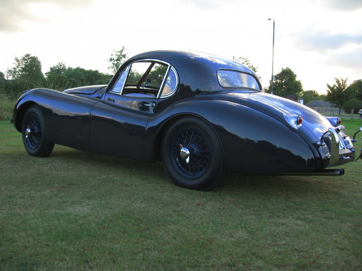 Fixed Coupe Pistonheads Head - The image features a classic black race car parked on a grassy area under clear skies. The car boasts a sleek, streamlined design with curved lines, particularly prominent around the wheel arches. It has chrome accents such as the rear fender, which add a vintage touch to the vehicle. The lush green grass contrasts with the stark black of the car, creating a striking visual. The setting gives the impression of the car being displayed at an outdoor event or exhibition.