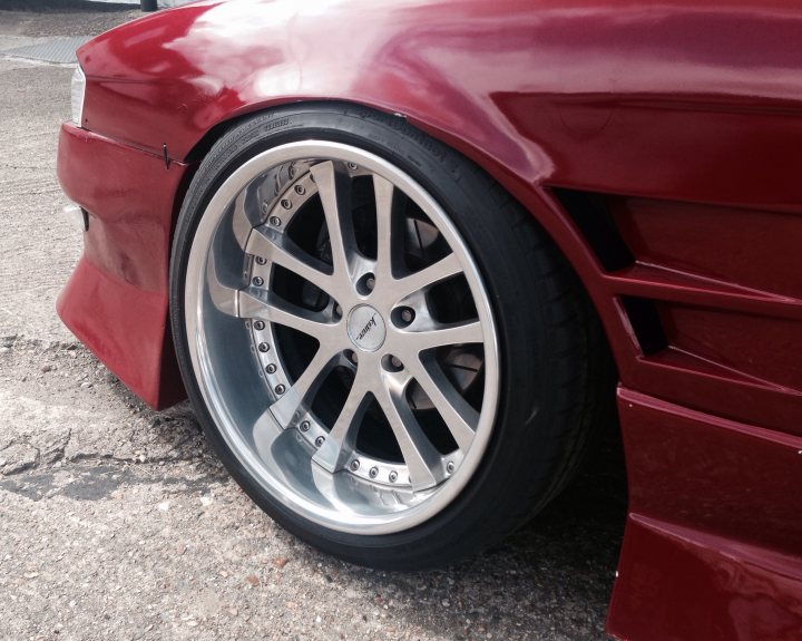 Miura SV being restored - Page 1 - Lamborghini Classics - PistonHeads - The image focuses on a close-up of the front wheel of a red car. The wheel exhibits a shiny chrome finish. The car is parked on a paved surface with gravel visible around it. In the background, there are some indistinct objects that suggest an urban environment. The overall scene conveys a sense of stillness in an otherwise bustling cityscape.