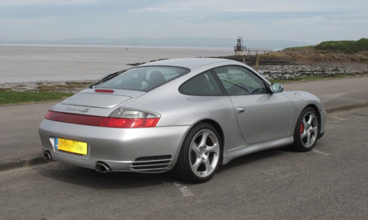 The image shows a silver Porsche Cayman sports car parked on a paved area next to a body of water. The car is positioned facing the camera towards the water, with a clear view of its sleek design and the distinctive red light strip along the back of its rear wings. The surrounding environment consists of a calm waterfront, with grassy areas next to the water and a less busy road extending into the distance. The car is presumably at rest with no immediate signs of motion. The image captures the car's elegant design, set against a tranquil natural landscape, suggesting a serene and picturesque setting.