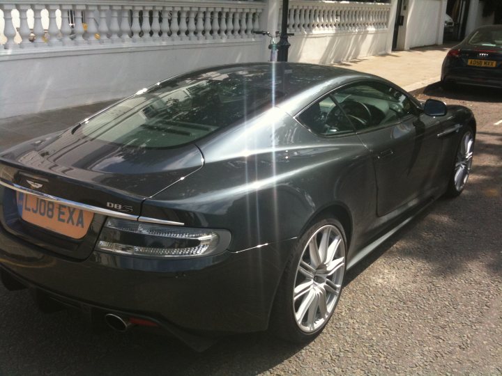 Any photo's of Aston's in  Tempest Blue - Page 1 - Aston Martin - PistonHeads - The image presents a scene featuring a black Audi R8 sports car on a street. The car is positioned perpendicular to the camera, showcasing its sleek silhouette. The hood of the car appears to be reflecting sunlight. In the background, there is a white gate with ornamental posts and a green lawn extending into the distance. Safety features, such as a license plate and side mirrors, are visible on the car, indicating it is a functional and legally registered vehicle. The setting suggests a suburban or rural residential area. A second, smaller car is visible in the far background on the right, indicating that this is a public roadway. The image captures a moment on the road, possibly during daylight.