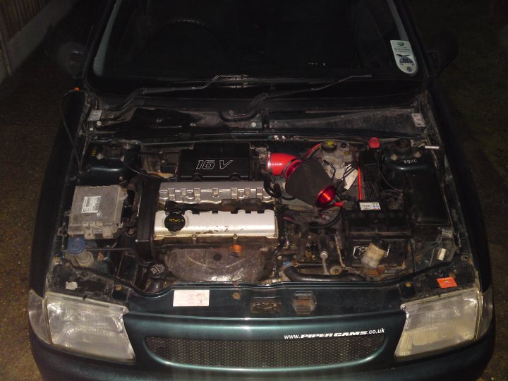 Pistonheads Sleeper Saxo - The image shows the hood of a car that has been taken off. The engine is exposed and there are various mechanical parts visible, with the motor being the most prominent. Above the motor, there's a red part with wires connected to it, possibly related to electrical components or systems of the vehicle. The sides of the hood are slightly bent, indicating the car has been recently repaired or maintained. There is a label or sticker with text visible near the front of the hood, but the text is not clearly legible. The background is dark and three black handles are visible, which may be part of the car's structure or access points.