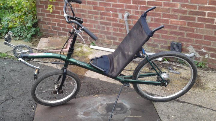 Pistonheads - This image features a heavily modified bicycle with several adaptive components. The bicycle has a long horizontal frame that supports an extended seat, creating unique seating options. To one side, there's a vertical trimmed, cushioned seat, which appears to have been removed or partially lifted off the bike. Nearby, there is a stack of old tires and other debris indicating a makeshift repair area or a pile of items in need of attention. The backdrop consists of a brick wall and green plants, suggesting this picture might have been taken in a backyard or similar outdoor space.