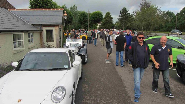 Hare Breakfast Meet - August - Page 1 - Kent & Essex - PistonHeads - This image depicts a group of people walking along a road next to parked cars, including a vintage white car and a green one. The people are casually dressed and some are carrying items like handbags or backpacks. In the background, there is a line of parked cars, and the sky is overcast, suggesting it might be a cloudy day. The scene appears to be a regular day on a busy street.