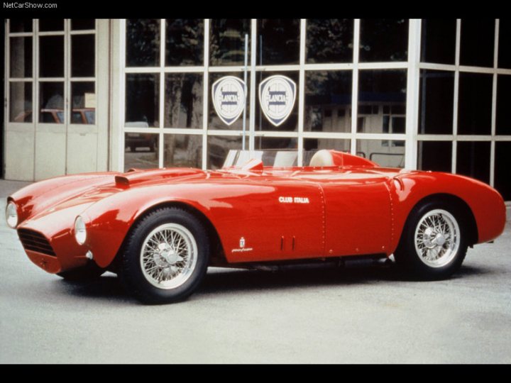 My Dilemma. A Retro TVR? - The image is a vintage photograph featuring a bright red sports car, which appears to be a veteran racer, as indicated by the distinctive shape and small size. It is parked in front of a building with large windows, which show up in the reflection of the car. The car features chrome details, including accents on the bodywork and silver rims. The tail of the car is green and displays the word "Panatucci" with an emblem above it. The style of the car suggests it might be from the early to mid-20th century, and the image itself has faded over time, adding to its historical feel.