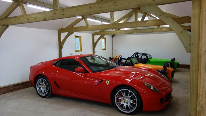 Who has the best Garage on Pistonheads???? - Page 132 - General Gassing - PistonHeads - The image shows a large garage where various racecars are parked. The racecars are sleek, with aerodynamic bodywork, and they are predominantly red and black in color. Some cars are fully exposed, while others have parts covered with black tarps. The garage has a high ceiling supported by exposed wooden beams, giving it a robust and industrial appearance. There are no people visible in the image.