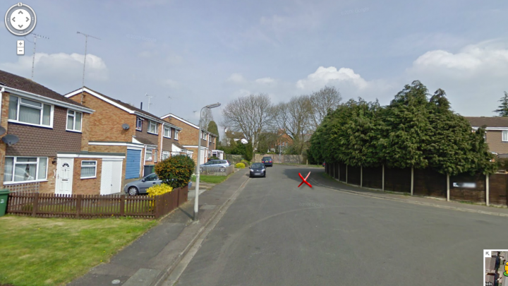 Legally what is a junction? Re: parking - Page 1 - Speed, Plod & the Law - PistonHeads - The image shows a quiet residential street. On the side of the road, there is a small grassy area with a few vehicles parked, and fences along the sidewalk. Across the street, there are houses with brick walls and white roofs. A tree with red leaves is prominently visible in the front yard of one of the houses. A closer look at the lower middle part of the image reveals a bright red "Do Not Enter" sign. On the bottom right corner, there seems to be an icon indicating an error or a pause in the video feed.