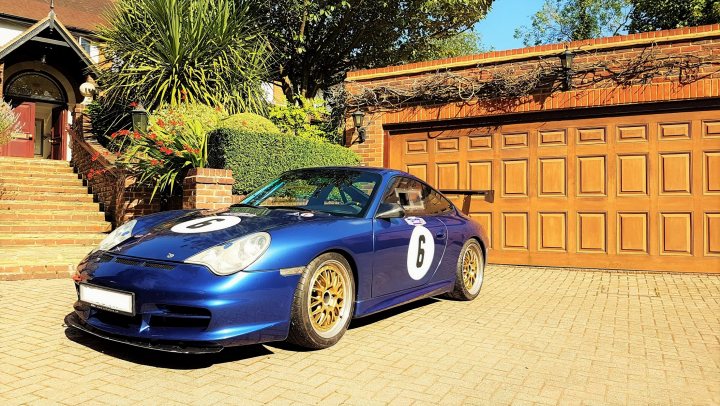 Is the bubble about to burst? - Page 152 - Porsche General - PistonHeads - The image showcases a blue Porsche 996 racing car parked in front of a garage. The car is painted in a glossy blue hue and exhibits notable racing accents such as the number six on the hood and various decals. Its hood is open, revealing a glimpse of its powerful engine. The garage has a door with a grid of wooden panels, and the car is positioned on a brick driveway.