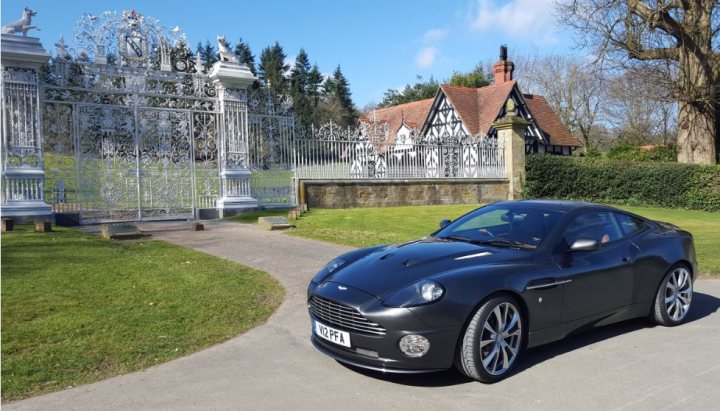 RE: Aston Martin Vanquish: Spotted - Page 2 - General Gassing - PistonHeads - The image features a black sports car parked on a road, with an ornate fence in the background. The vehicle appears to be a convertible and is positioned to the left of the frame. There are no visible people in the image, focusing attention on the car and its surroundings. The setting suggests a well-maintained area, possibly a park or a similar public space, given the presence of the fence and the paved road. The lighting indicates it could be midday, as shadows are not overly prominent.