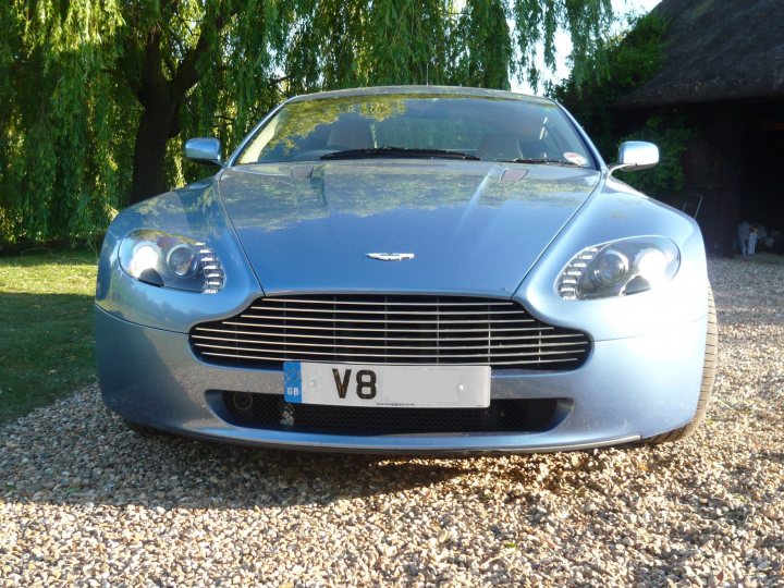 V8V Newbie (ish) - Page 1 - Aston Martin - PistonHeads - The image features a silver Aston Martin car parked in a driveway. The vehicle appears to be a V8 model, as indicated by the license plate that reads "V8." The Aston Martin emblem is prominently displayed on the front grille. The car is sleek and polished, reflecting some of the surrounding elements in its shiny surface. The setting is outdoors, with a gravel driveway visible beneath the car and a garage in the background. The natural environment includes trees and grass, suggesting a serene residential area.