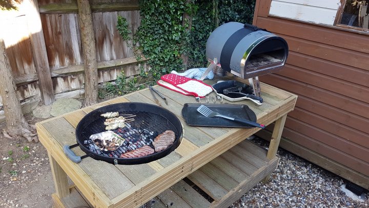 Pallet wood Weber grill table project on a budget - Page 3 - Homes, Gardens and DIY - PistonHeads - The image portrays an outdoor cooking scene. In the center is a black, shell-shaped grill with pieces of meat sizzling on it. To the left of the grill, there's a rectangular black tray with a grill tool and what appears to be foil on it. A red cloth is unrolled on the left side of the grill with its back towards us. In the background, there's a brown wooden structure to the right and a green tree in the corner behind the BBQ. The outdoor setting is indicated by the natural light and the structure's siding. The focus and composition of the photo center on the grill as the main subject.