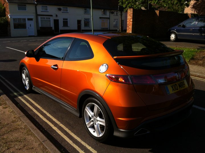 Orange Cars - Page 14 - General Gassing - PistonHeads - The image shows a brightly daylit scene. An orange hatchback car is parked near the curb, with its back to the viewer. It features a round badge and a license plate on the back. In the background, there are residential houses and a path. The photo has a clear color contrast, with the car highlighting the street's grass and shrubbery.