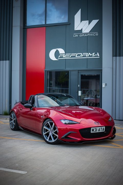 2016 Mx5 Recaro - many modifications  - Page 1 - Readers' Cars - PistonHeads - The image features a vibrant red sports car parked in front of a building with a sign that reads "WORLD GARAGE". The car is positioned on the left side of the frame, facing towards the right. Behind the vehicle, there is a contrasting green building with a large white letter 'C' on it. Above the car, there is a sign that says "LUXURY CARS", indicating the nature of the business at this location. The overall scene suggests a place dedicated to luxury vehicles, possibly a dealership or service center.