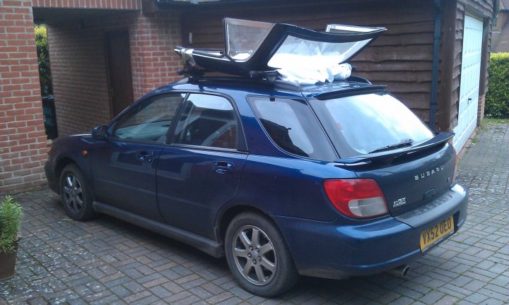 A car parked in front of a parking meter - Pistonheads - This image features a **blue Toyota SUV** parked in a driveway, partially covered by an **ultraviolet sunshade**. The SUV is a **Suzuki**, as indicated by the visible model name. The car is parked in front of a building with **brick walls** and a **skylight**. The driveway appears to be made of **tarmac**. There is a slight reflection on the car, suggesting wet or dewy conditions.