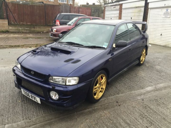 A car is parked on the side of the road - Pistonheads - The image showcases a dark blue sports car with a prominent air intake on the hood, parked on a concrete surface. The vehicle is positioned in front of a white garage door, which is partially obscured by the car. Behind the car, on the left side, there's a dark-colored vehicle, possibly an SUV. The sky appears overcast, suggesting it might be a cloudy day.