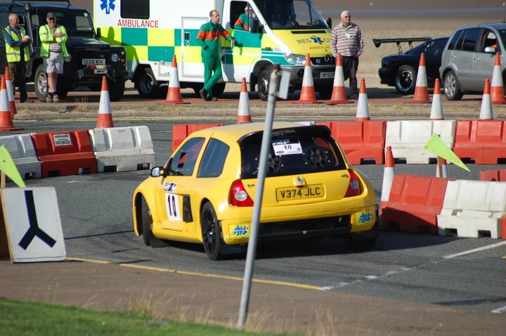 A yellow taxi cab parked in front of a building - Pistonheads - The image is a lively scene at a motorsport event. Dominating the foreground is a vibrant yellow rally car labeled with the number 10. The rally car is in motion, weaving through barriers painted in alternating red and white stripes. The backdrop is a bit busier, featuring a mix of emergency vehicles, including an ambulance and a police car, and other spectators, adding to the atmosphere of the event.