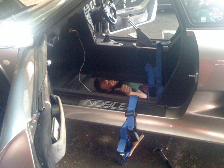 Floor reseal idea - Page 1 - Noble - PistonHeads - The image depicts a person, presumably a woman given the firmness of her posture and the visible hairstyle, lying reclined in the trunk of a car. A car seat is secured by blue seat belts. The trunk is open, suggesting that the vehicle is in a servicing environment or the photo is taken after the car seat has been removed for a creation or repair activity. The partially visible car parts and the presence of another similar car in front of the first one further support the idea that this is a workshop setting. The scene is lit with artificial lighting, contributing to the functional and utilitarian atmosphere.