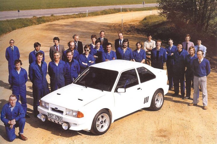 Pistonheads Market Close - The image is a group photograph featuring a team of individuals dressed in matching blue jumpsuits. They are standing in front of a white rally or racing car, which is positioned on a dirt surface. The setting includes a clear sky above and open fields in the background, with the horizon visible in the top left corner. The team members appear to be standing in a line, with most of them looking towards the camera, exuding a sense of unity and purpose. The positioning of the team and the car gives a dynamic feel to the photograph.